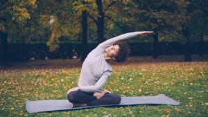 Slim young lady is doing sequence of yoga asanas sitting on mat in park alone on warm autumn day with beautiful grass and trees around her. Recreation and sport concept.